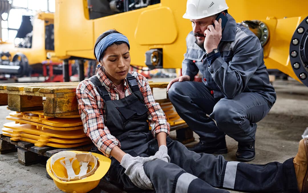 oung female engineer with hurting knee sitting on the floor by anxious male worker in safety helmet and uniform calling ambulance